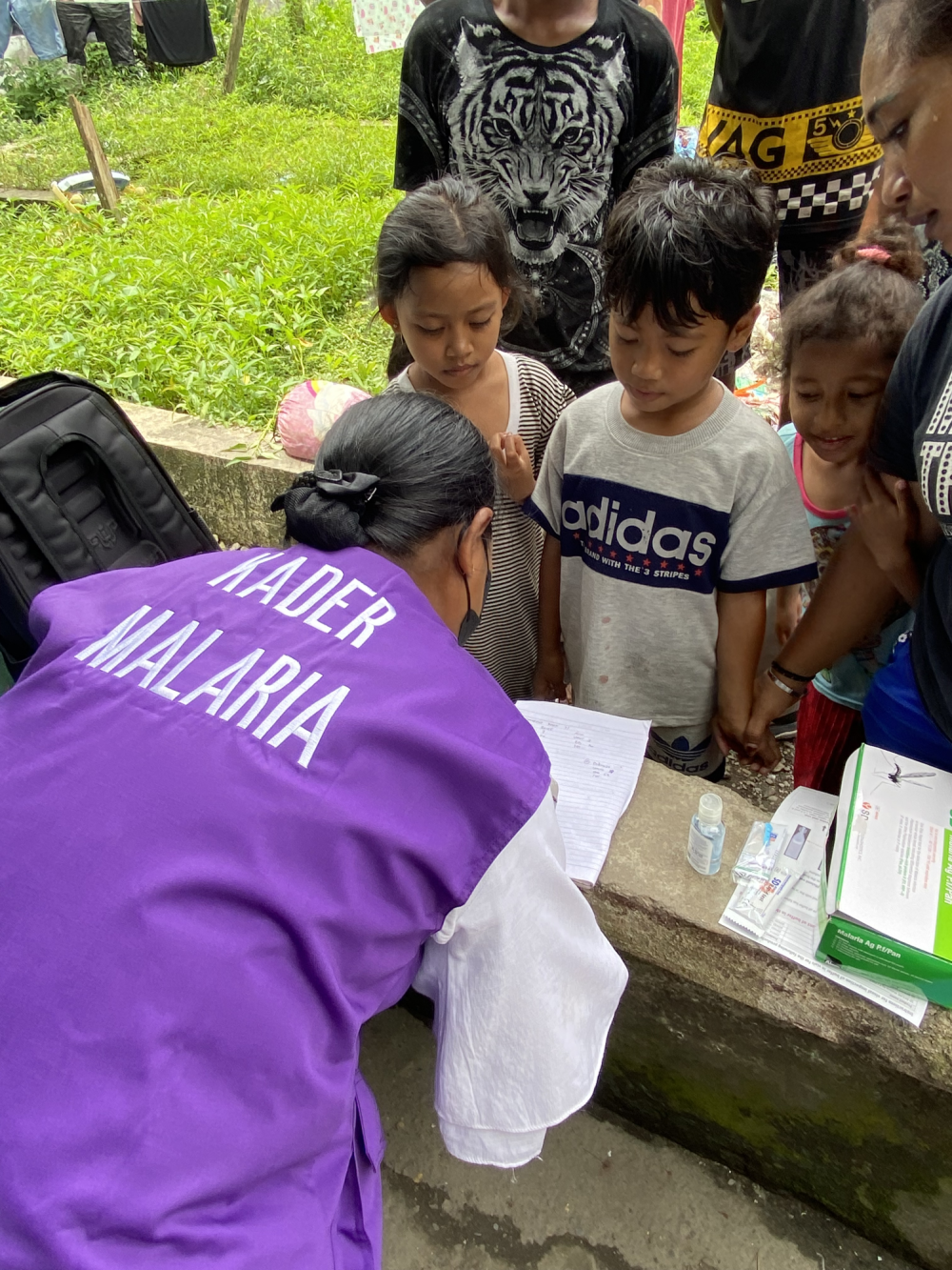 A female malaria cadre in purple vest working with children.